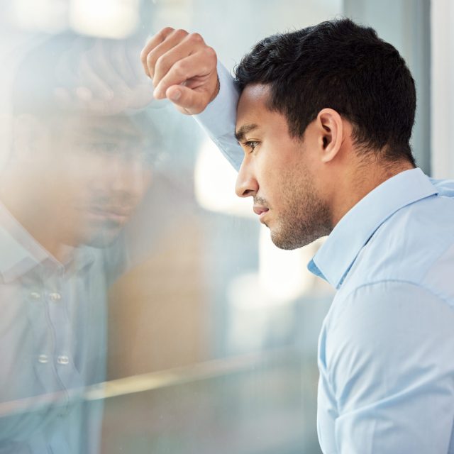 Shot of a young businessman leaning against a window looking depressed.