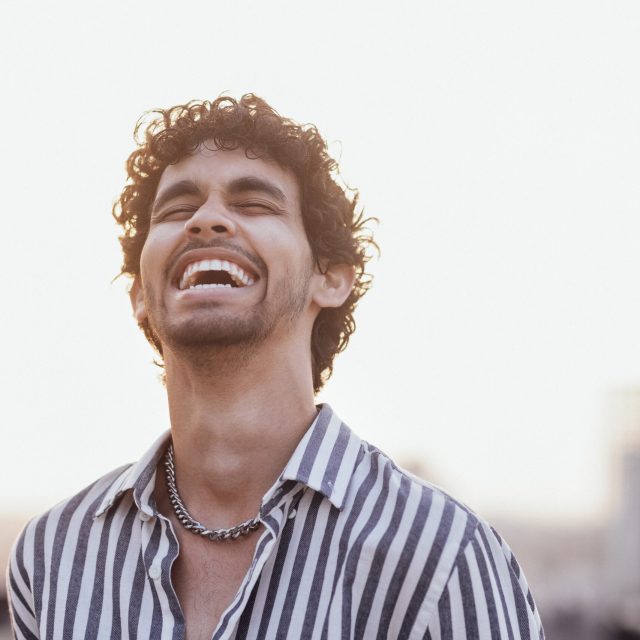 Close-up of a male portrait of a young man of mixed race. An attractive curly-haired guy in a striped shirt smiles against the backdrop of a big city. Cute laughing human on the roof of the house.