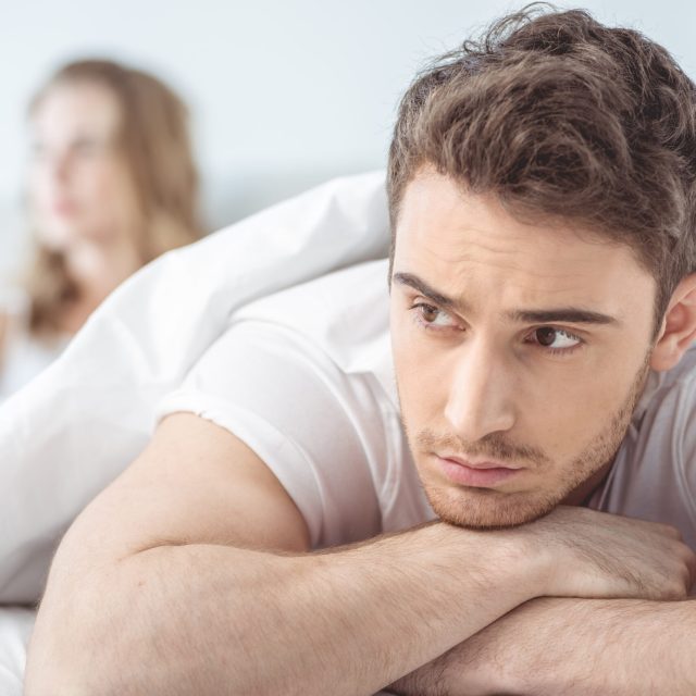 young dissatisfied man lying in bed with girlfriend behind