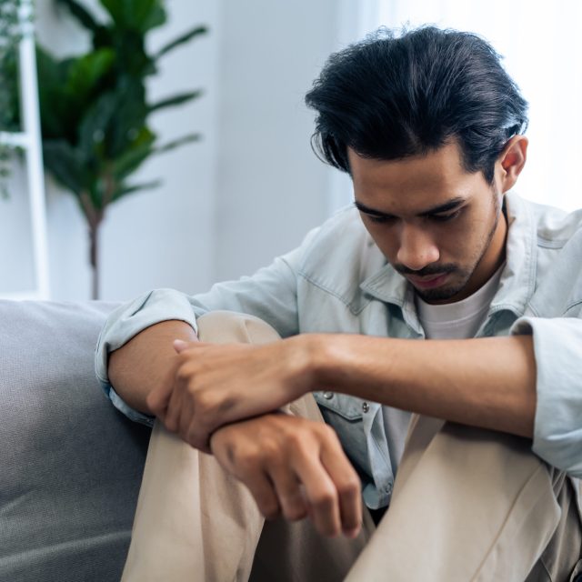 Asian young upset depressed man sitting alone in living room at home. Attractive unhappy male feeling very sad, lonely and frustrated with life problem think of money debt, budget loss, bankruptcy.