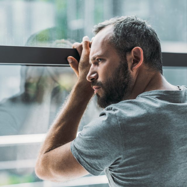 upset bearded man leaning at window and looking through it