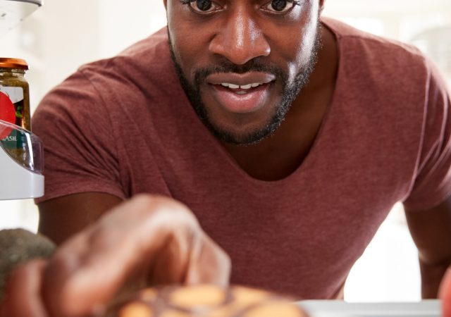 View Looking Out From Inside Of Refrigerator As Man Opens Door And Reaches For Unhealthy Donut